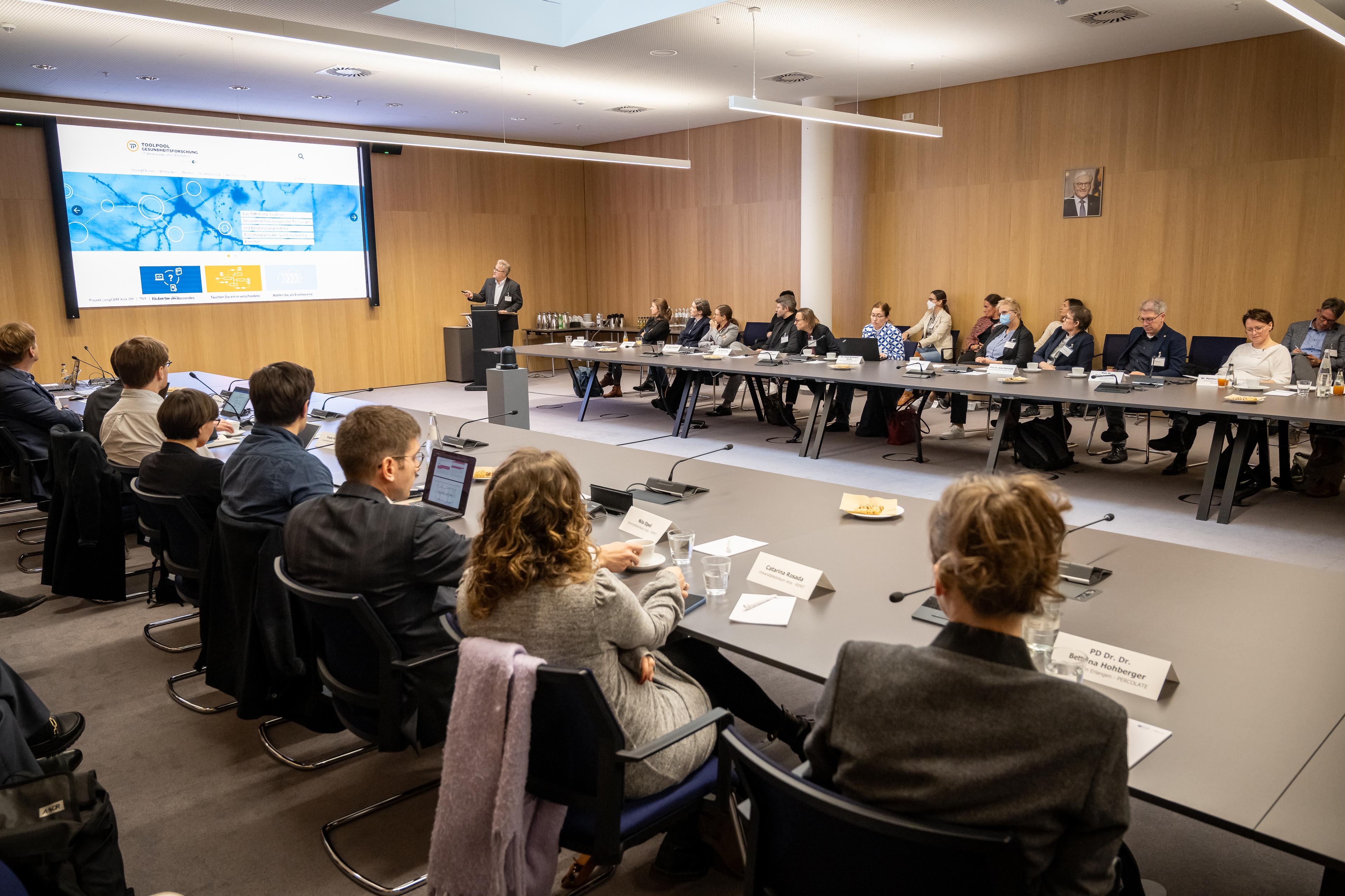 Ein Konferenzraum mit einer großen rechteckigen Tischanordnung, an der zahlreiche Personen sitzen. Ein Redner steht vorne neben einer Leinwand und hält eine Präsentation mit einer Folie, die Informationen zum Forschungsnetzwerk Long COVID zeigt.