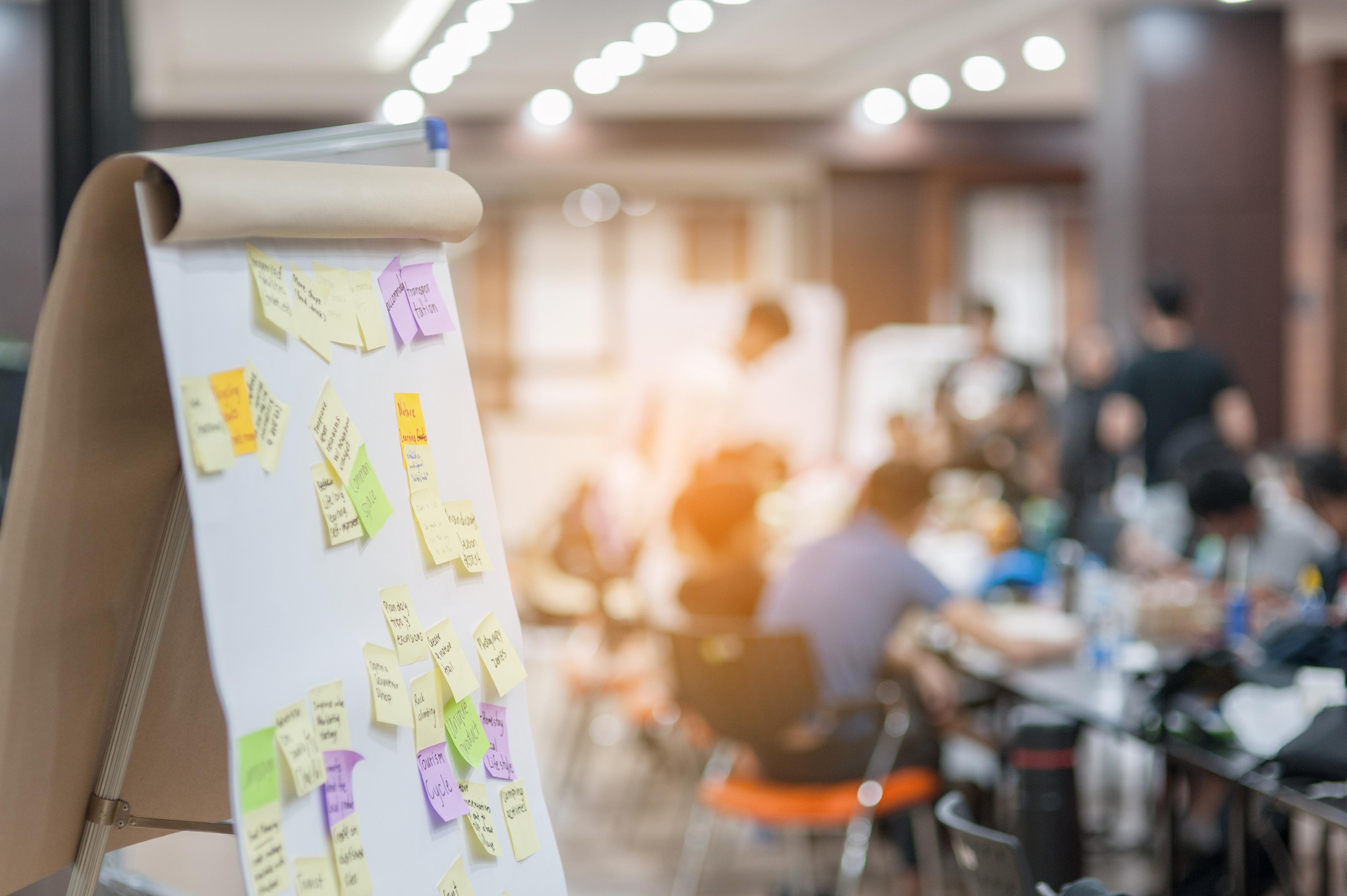 Das Foto zeigt ein Flipchart mit mehreren Notizen. Im Hintergrund mehrere Personen zu sehen, die alle um einen Tisch herum verteilt sind und gemeinsam arbeiten.
