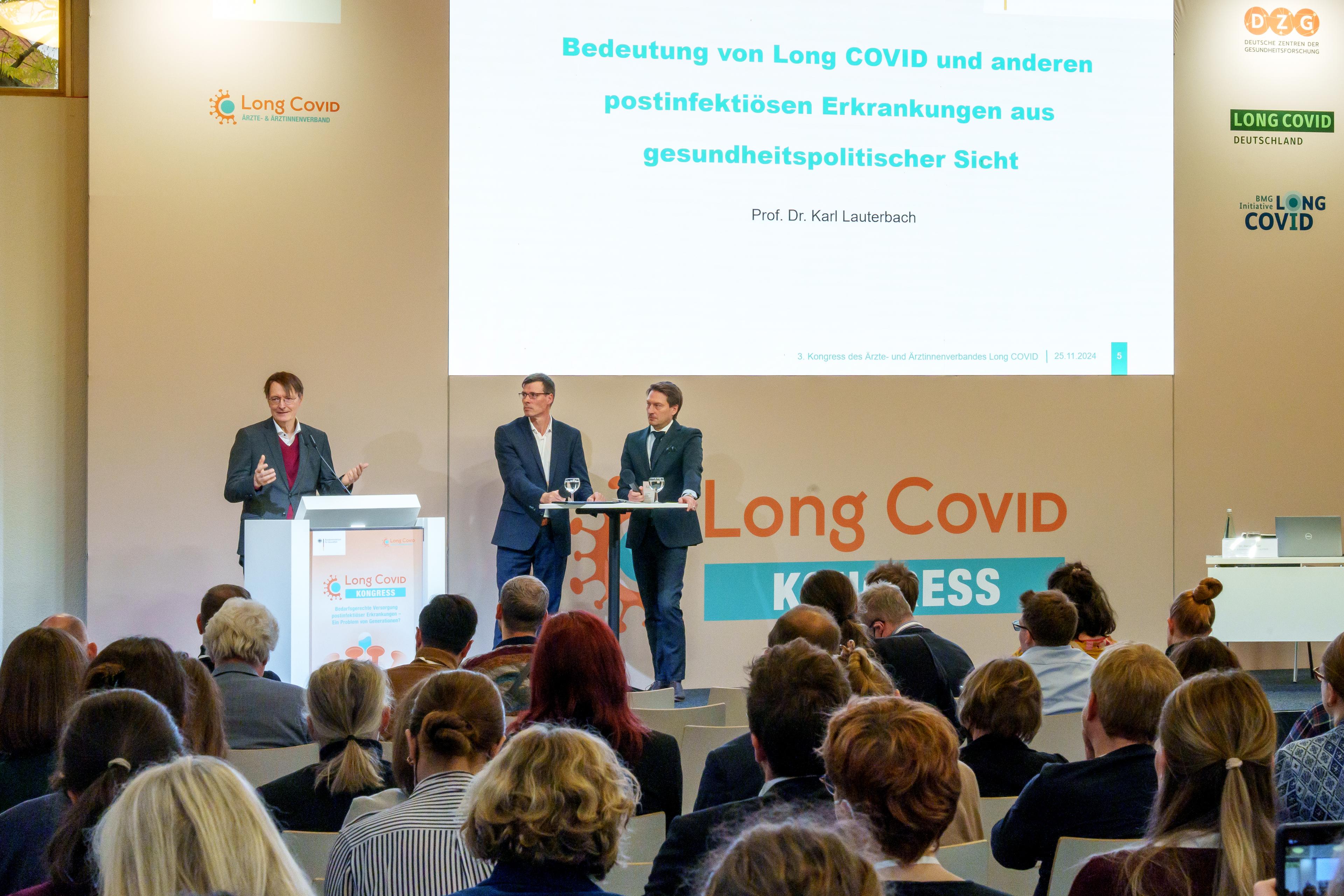 Bundesgesundheitsminister Prof. Dr. Karl Lauterbach spricht am Rednerpult bei der Eröffnung des 3. Long-COVID-Kongresses in Berlin. Im Hintergrund ist eine Leinwand mit dem Titel seines Vortrags zu sehen: "Bedeutung von Long COVID und anderen postinfektiösen Erkrankungen aus gesundheitspolitischer Sicht". Neben ihm stehen zwei weitere Personen an einem Podiumstisch, während die Zuhörerinnen und Zuhörer aufmerksam folgen.