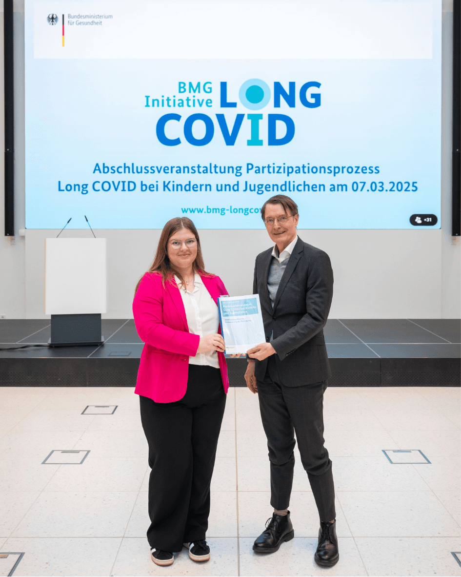 Bundesgesundheitsminister Prof. Dr. Karl Lauterbach und eine Betroffenenbotschafterin des Partizipationsprojekts „Long COVID bei Kindern und Jugendlichen“ stehen gemeinsam vor einer Leinwand mit dem Logo der Initiative. Die Betroffenenbotschafterin überreicht dem Minister ein Dokument mit Handlungsempfehlungen zur Verbesserung der Versorgung von Kindern und Jugendlichen mit Long COVID.